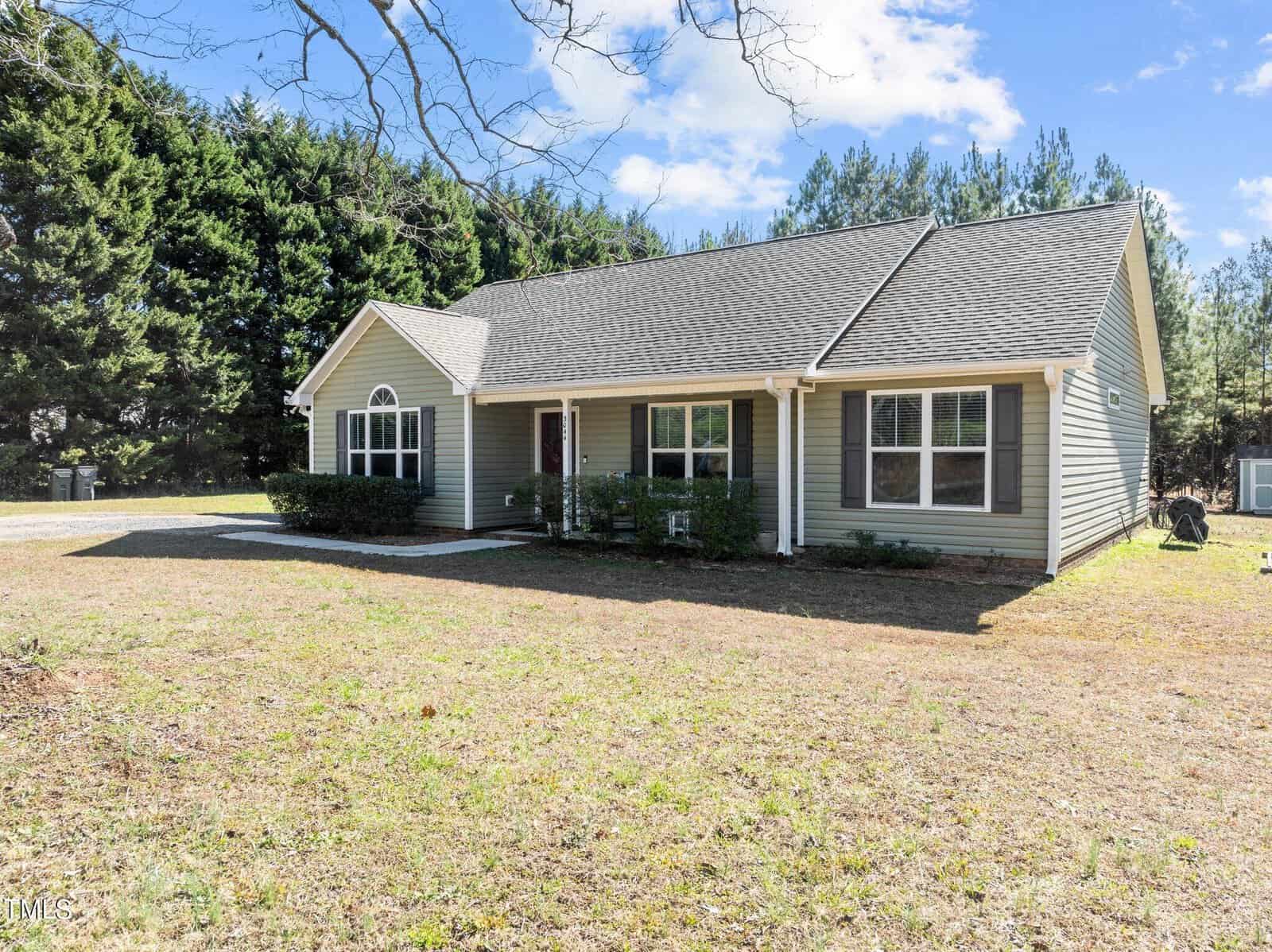 Single-story house with gray siding and a dark roof, surrounded by a grassy yard. Tall green trees line the background under a partly cloudy blue sky. A driveway is visible on the left side.