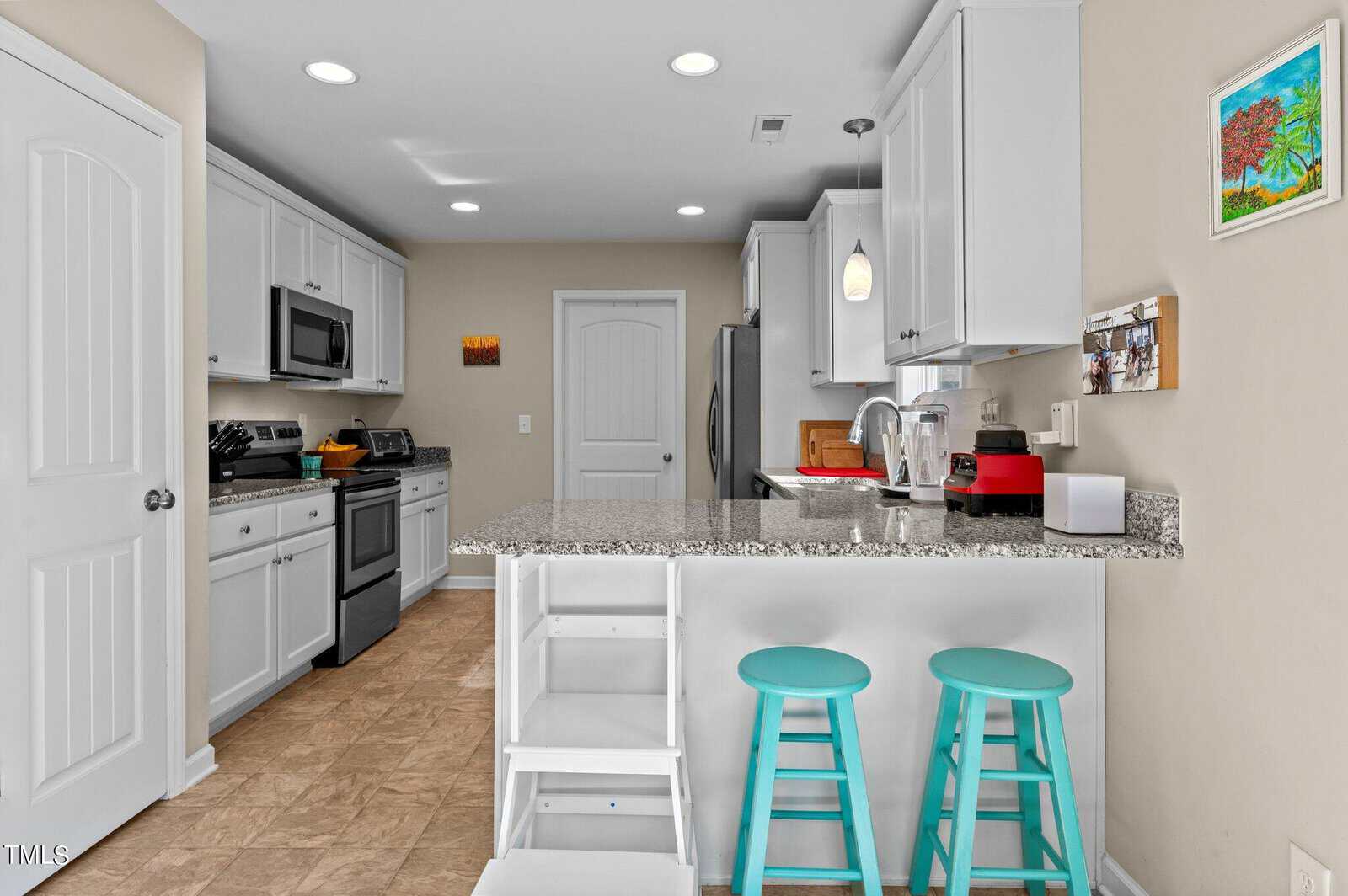 Modern kitchen with white cabinets and stainless steel appliances. A central island features a granite countertop with two turquoise stools. Art decorates the beige walls, and bright lighting enhances the clean, airy feel.