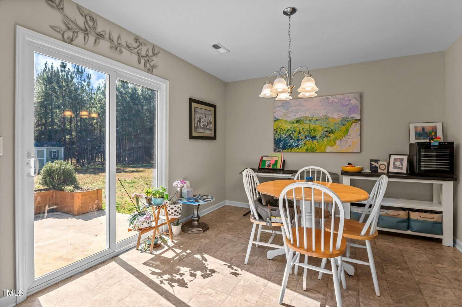 A dining area with a round wooden table and four white chairs. A colorful painting hangs on the wall. Sunlight streams in through a glass sliding door, revealing a view of a backyard with trees. A side table holds plants and decorative items.