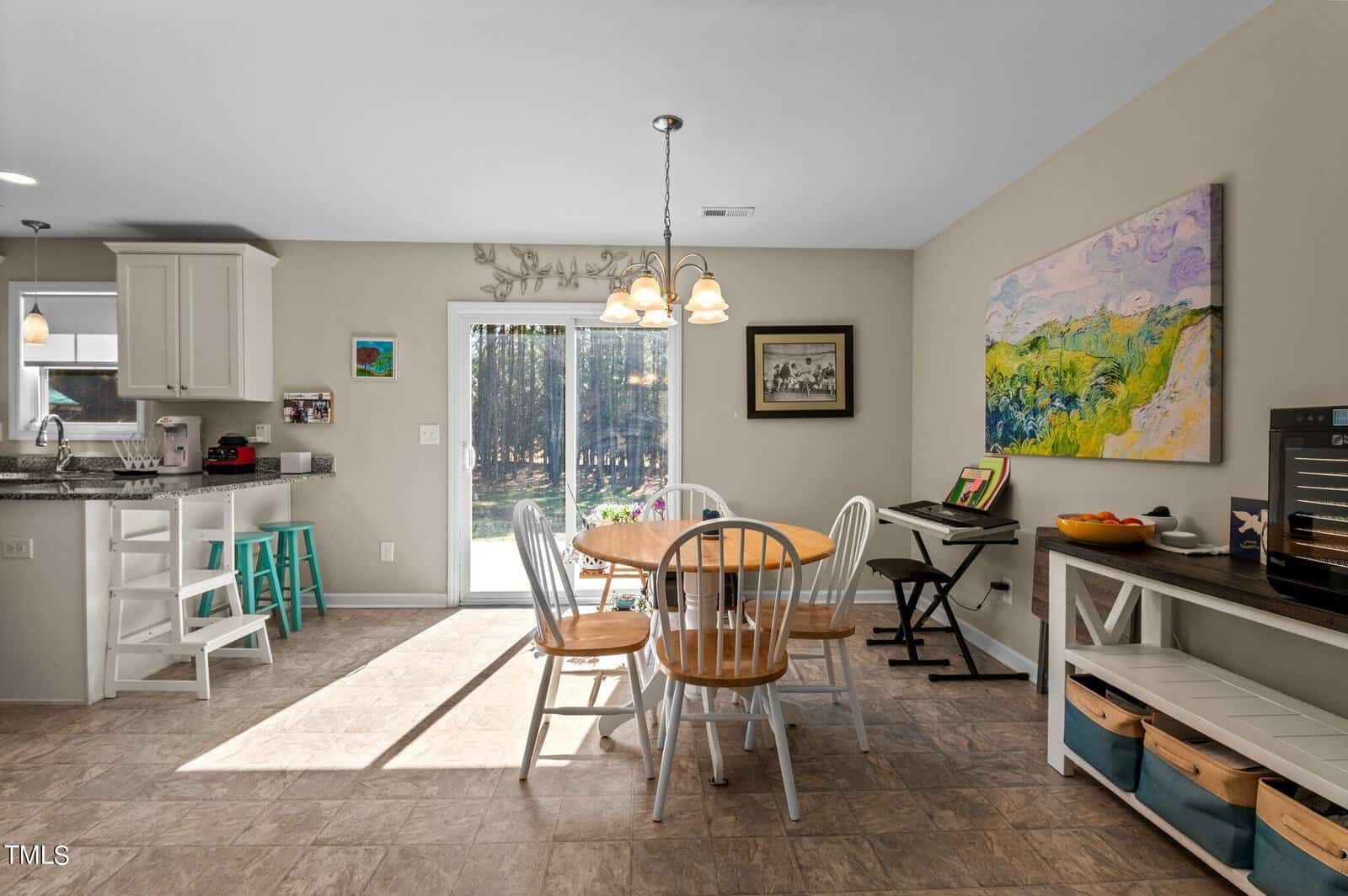 A bright kitchen and dining area with a round wooden table and chairs. The kitchen has white cabinets and a teal stool. Artwork is displayed on the walls. Sunlight streams through a sliding glass door, illuminating the tiled floor.