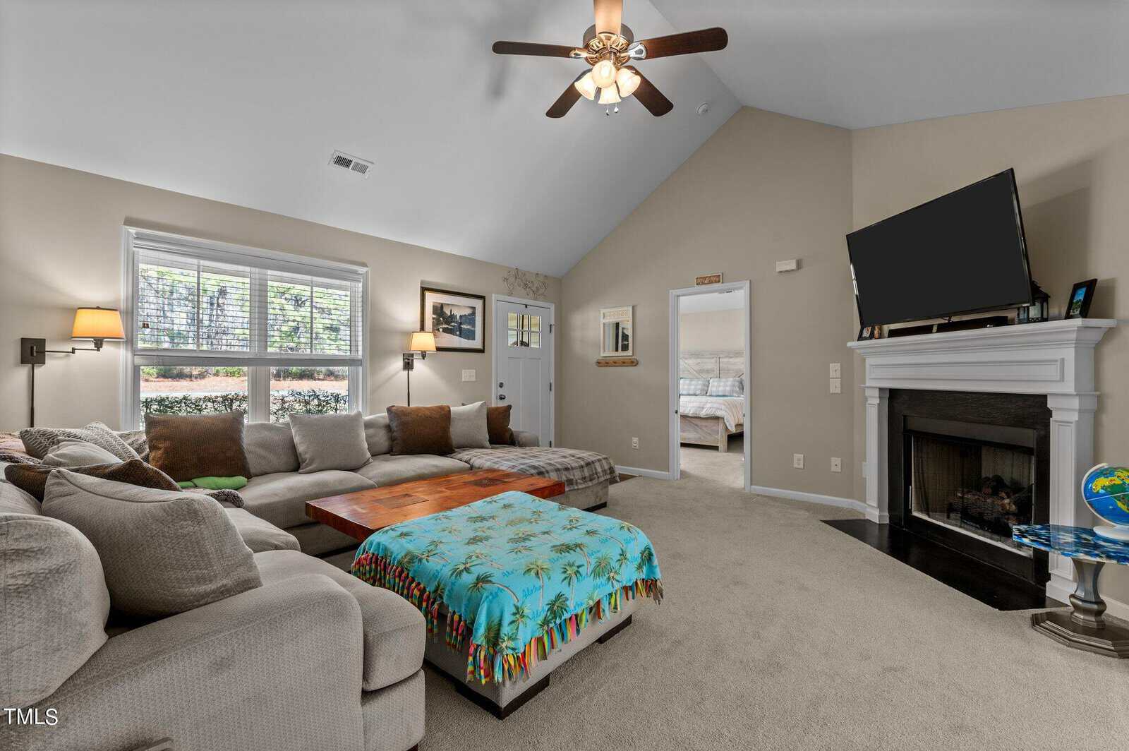 A cozy living room with a vaulted ceiling and fan. It features a large L-shaped sectional sofa with various cushions, a wooden coffee table, a fireplace, and a TV. The room is carpeted and has windows letting in natural light. A bedroom is visible through an open door.