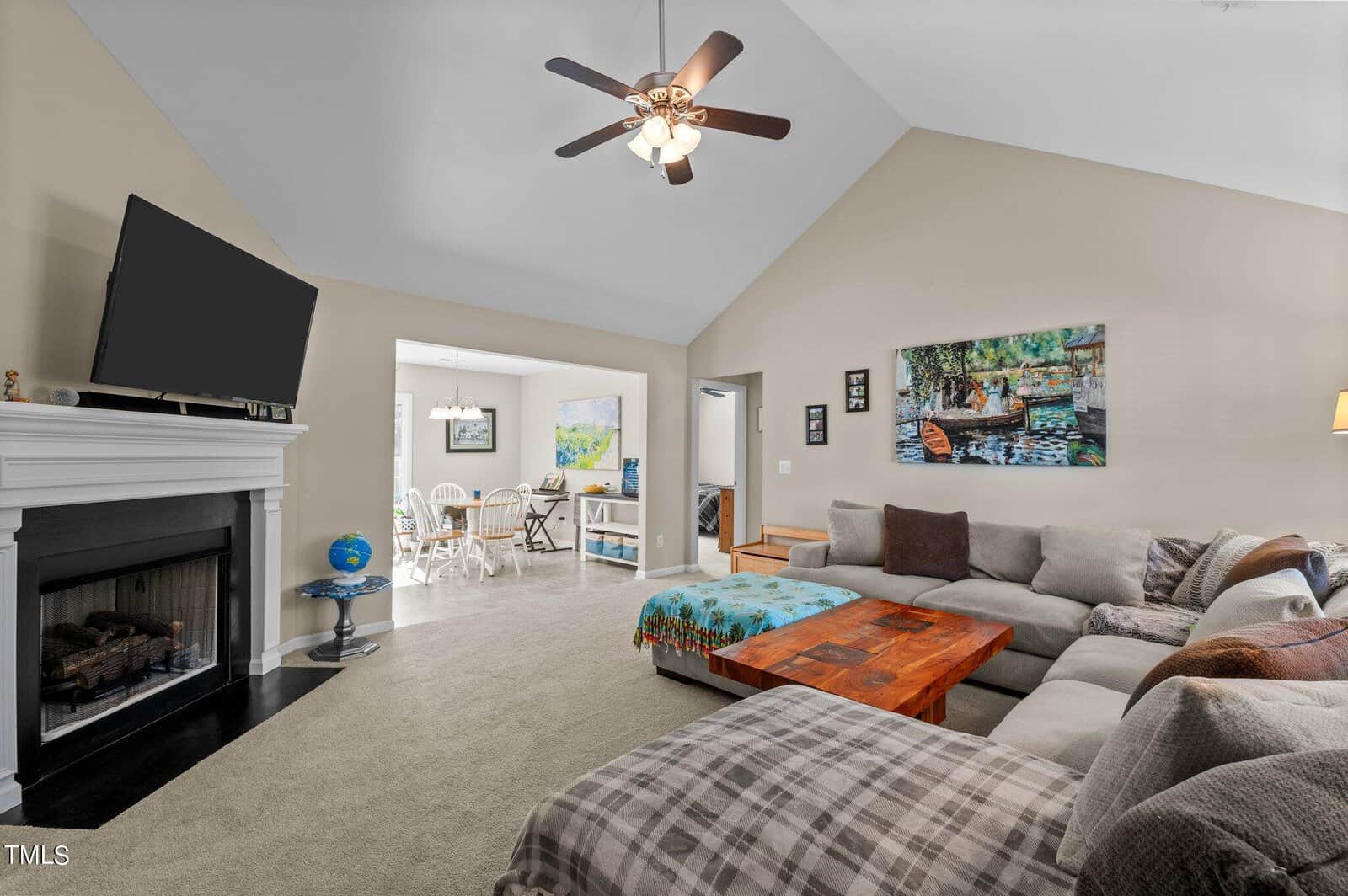 A spacious living room with a high ceiling and a ceiling fan. It features a sectional sofa with cushions and a wooden coffee table. A fireplace is on the left with a TV above it. In the background, a dining area and a wall with artwork are visible.