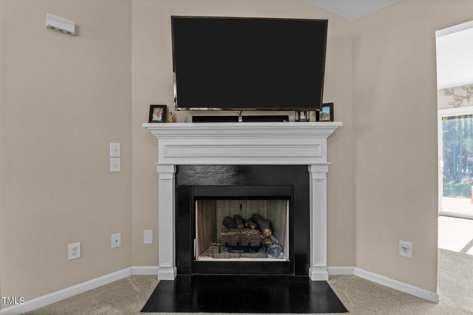 A living room corner featuring a white mantel fireplace with a black interior. Above the fireplace is a large, mounted flat-screen TV. Two small framed photos are on the mantel, and sunlight filters in from an adjacent window.