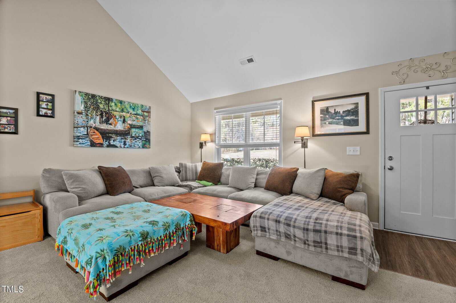 A cozy living room features a large, L-shaped gray sofa with various cushions, a wooden coffee table, and two ottomans covered with patterned throws. Two artworks hang on beige walls, and natural light streams through two windows near the door.