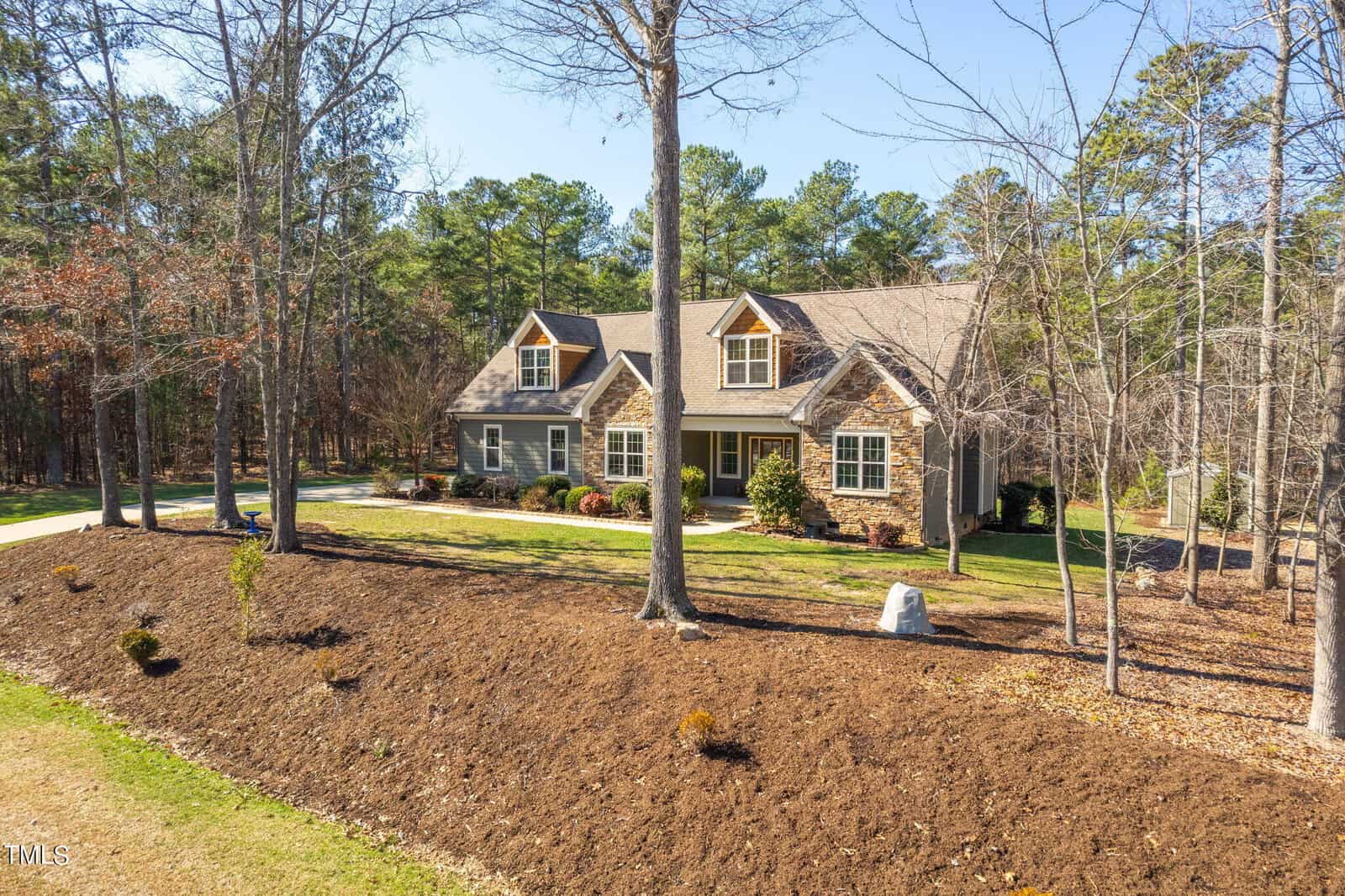 A charming two-story house with a gray exterior and a landscaped front yard. The home is surrounded by tall trees and set on a spacious lawn. A stone pathway leads to the front door, and the scene has a clear, sunny sky overhead.