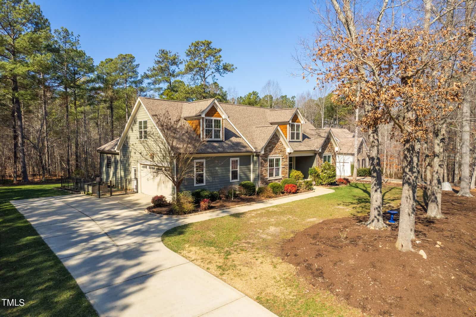 A spacious two-story house with a stone and green exterior sits amidst tall trees. The property features a neatly manicured lawn, a long driveway leading to a two-car garage, and a forested backdrop under a clear blue sky.