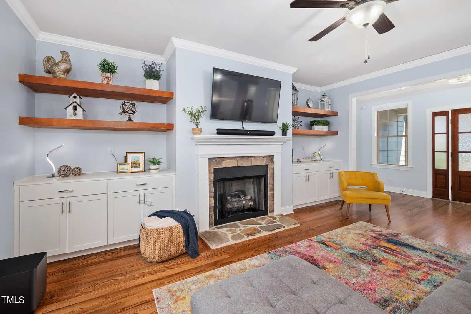 A cozy living room with light blue walls, a TV mounted above a fireplace, and wooden shelves on either side. There is a colorful rug, a yellow accent chair, and a gray tufted ottoman. Hardwood floors complete the inviting atmosphere.