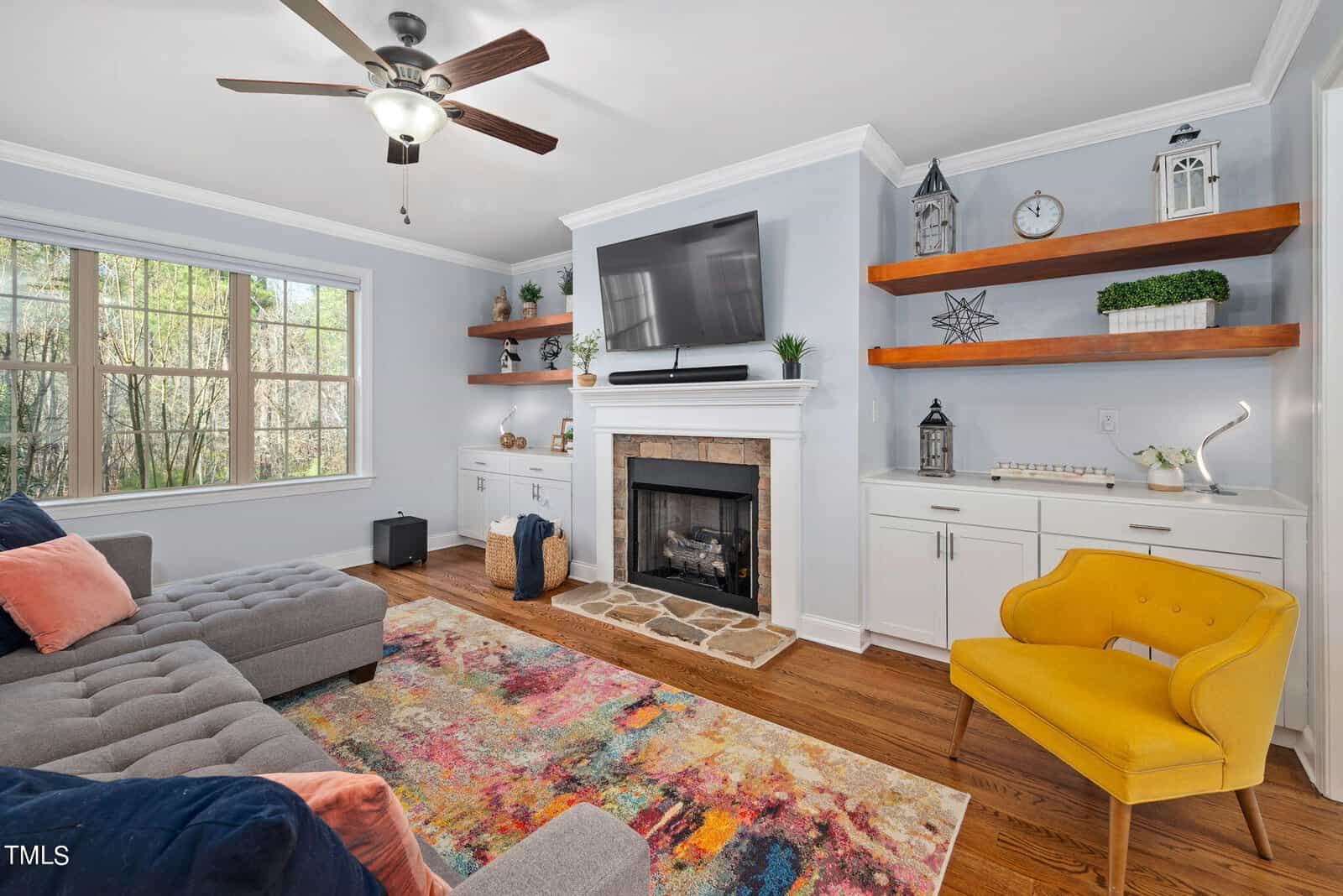 A cozy living room with a modern aesthetic. It features a gray sectional sofa, a yellow armchair, and a colorful rug. There's a fireplace with a TV above it, and wooden shelves with decorative items. Large window offers natural light.