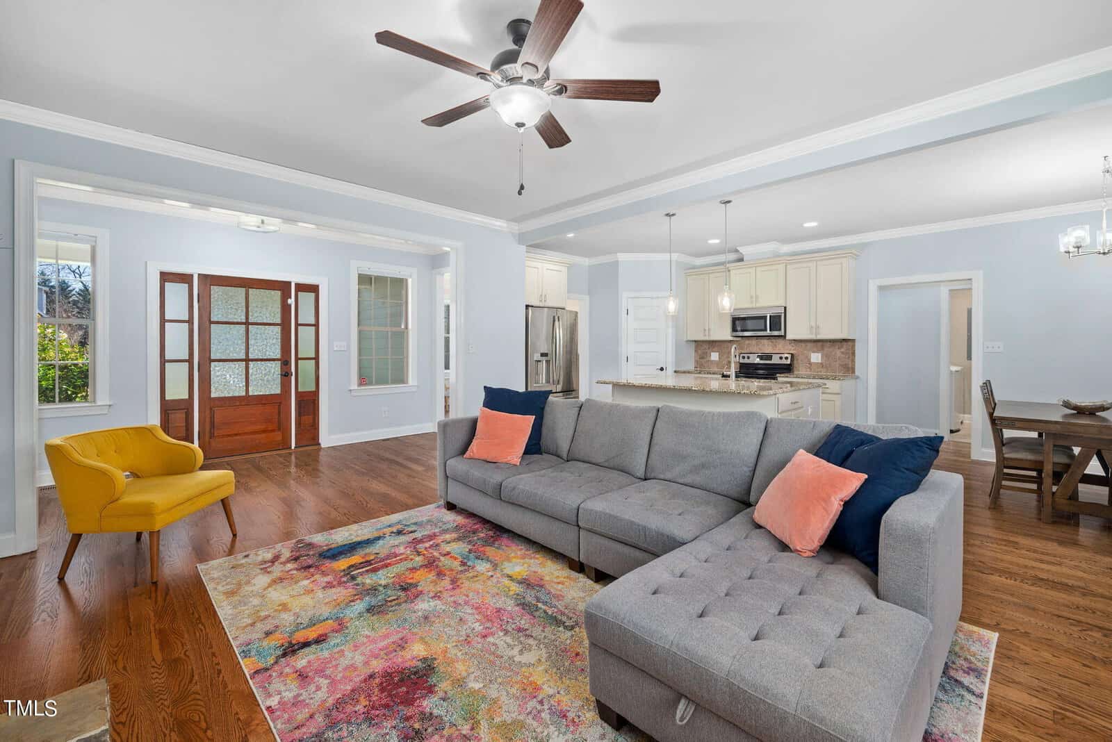 A bright living room with a gray sectional sofa featuring orange and navy pillows, a colorful area rug, and a yellow accent chair. The room opens into a white kitchen with modern appliances and a wooden dining table in the background.