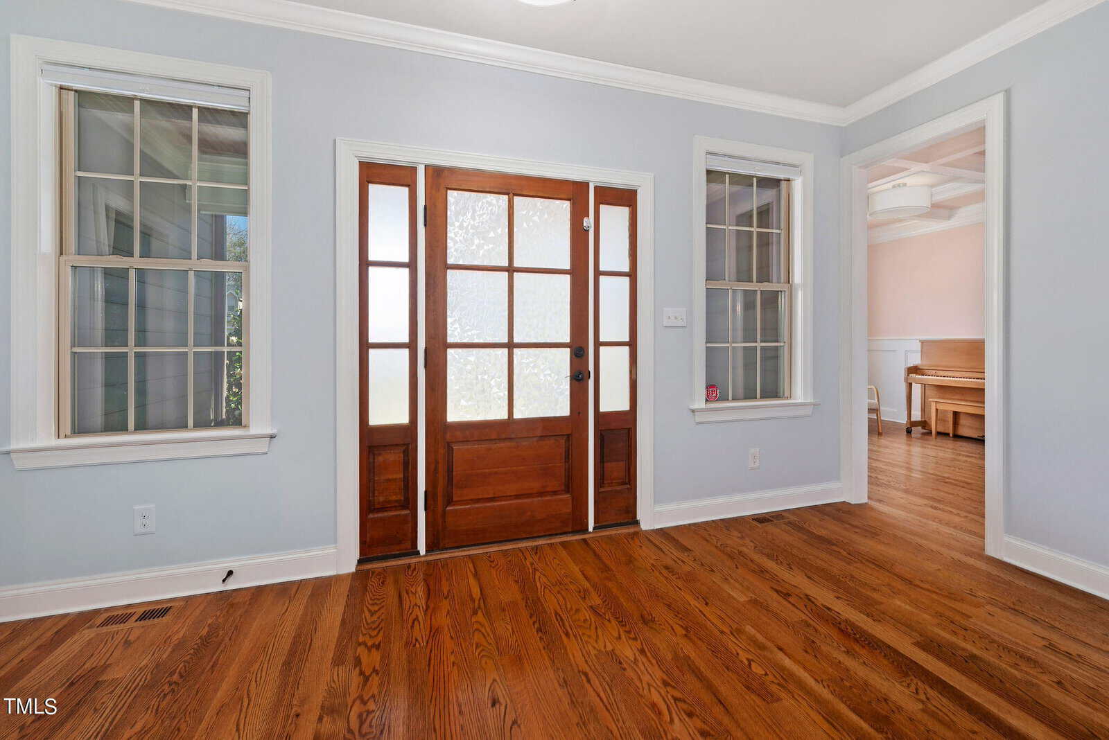 A room with light blue walls features a wooden floor and a symmetrical arrangement. A central wooden door with frosted glass panels is flanked by two windows, and there is a glimpse of a piano in the adjacent room.
