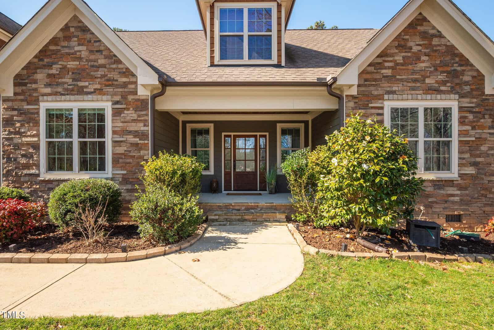 A charming house with a stone and wood exterior, featuring a covered front porch. The entrance is framed by neatly trimmed bushes and a small tree, with a concrete walkway leading to the front door. The house has a prominent dormer window.