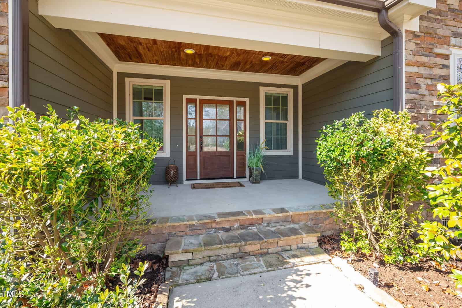 Front porch with a brown wooden door framed by two windows. Green siding and stone accents add character. Potted plants and shrubs line the walkway, creating a welcoming entrance. A wooden ceiling enhances the cozy atmosphere.