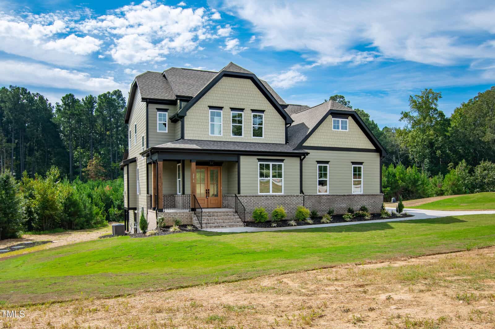 A two-story green house with a gray roof is set on a landscaped lawn. It has multiple windows and a wooden front door with stone steps leading up to it. Trees and shrubs surround the property under a partly cloudy sky.