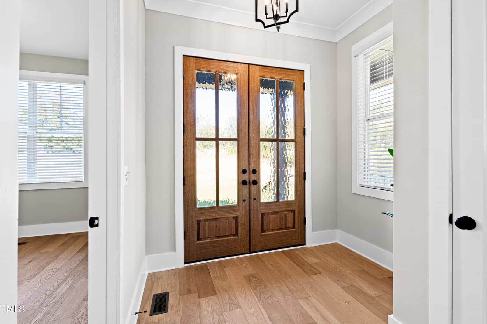 A bright entryway with double wooden doors featuring glass panels and black handles. The room has light-colored walls, a wooden floor, and a small window to the left with white blinds. A modern ceiling light fixture is visible above.