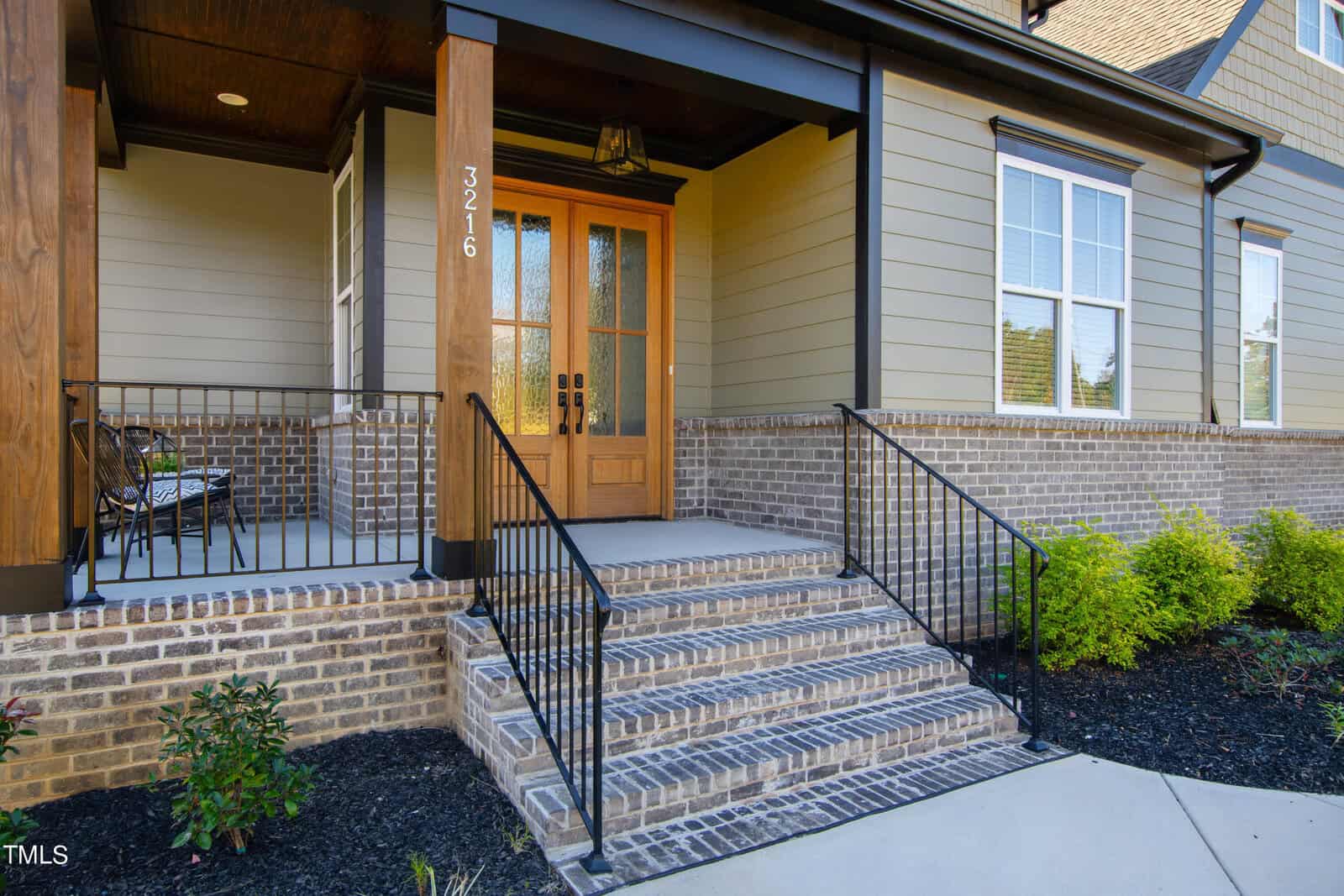Front entrance of a house with a wooden and glass door, brick steps, and a small porch. The exterior features beige siding and brickwork. Black railings line the steps and porch, with a small seating area to the left and shrubs along the front.