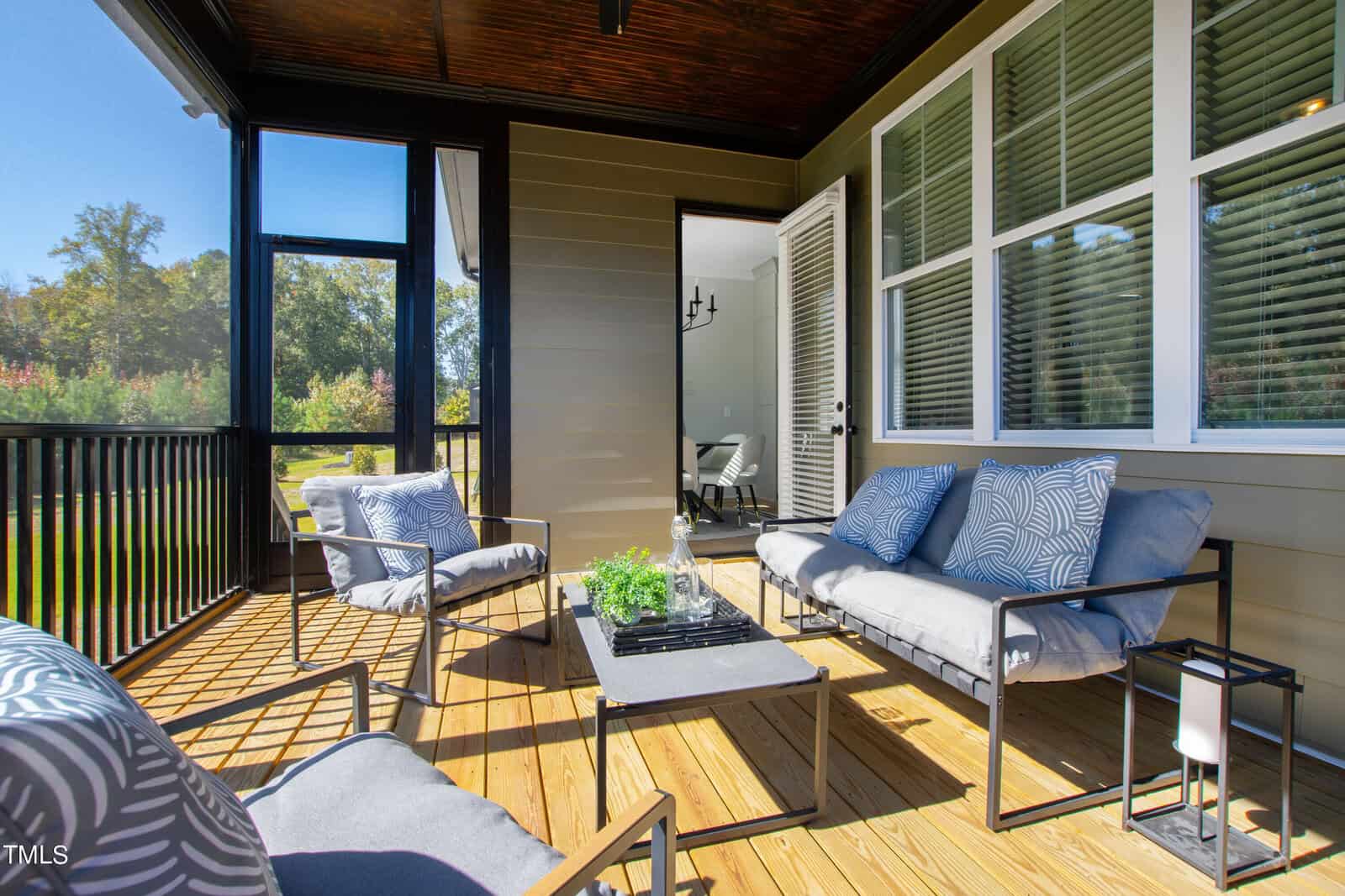 A cozy outdoor patio features a wooden floor, two cushioned chairs, and a sofa with patterned pillows. A coffee table with a small plant sits in the center. The area is enclosed by railing and is adjacent to a building with large windows and a glass door.