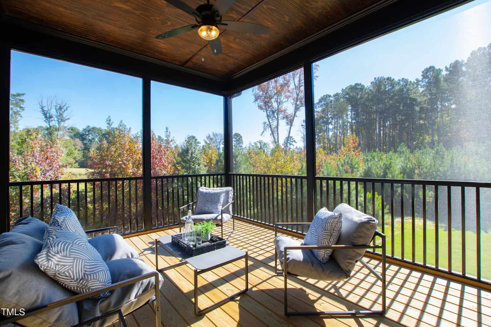 A screened-in porch with a wooden floor features a ceiling fan and three cushioned chairs around a low table. The space overlooks a sunny, scenic view of trees and a grassy area in the background.