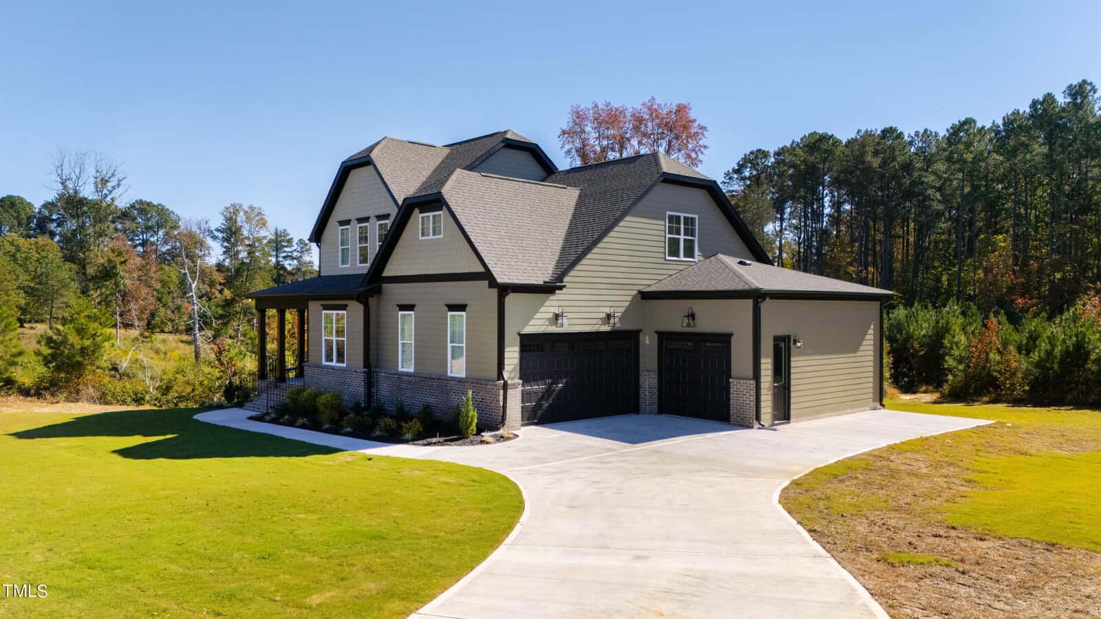 A two-story house with green siding and dark gray roof is situated on a spacious lot. It features a double garage and a paved driveway. Surrounding the house are green lawns and a backdrop of trees. The sky is clear and the weather appears sunny.