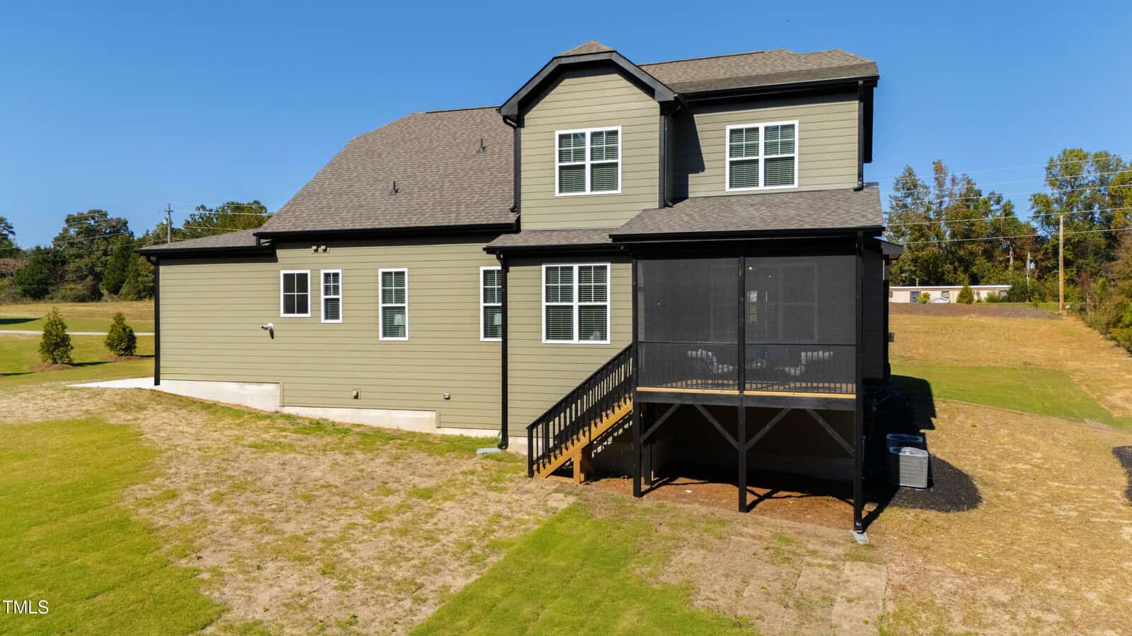 A two-story house with green siding is situated on a grassy lot. The house features a screened porch with a staircase leading to the yard and several windows on both floors. The surrounding area has sparse trees and clear blue sky.
