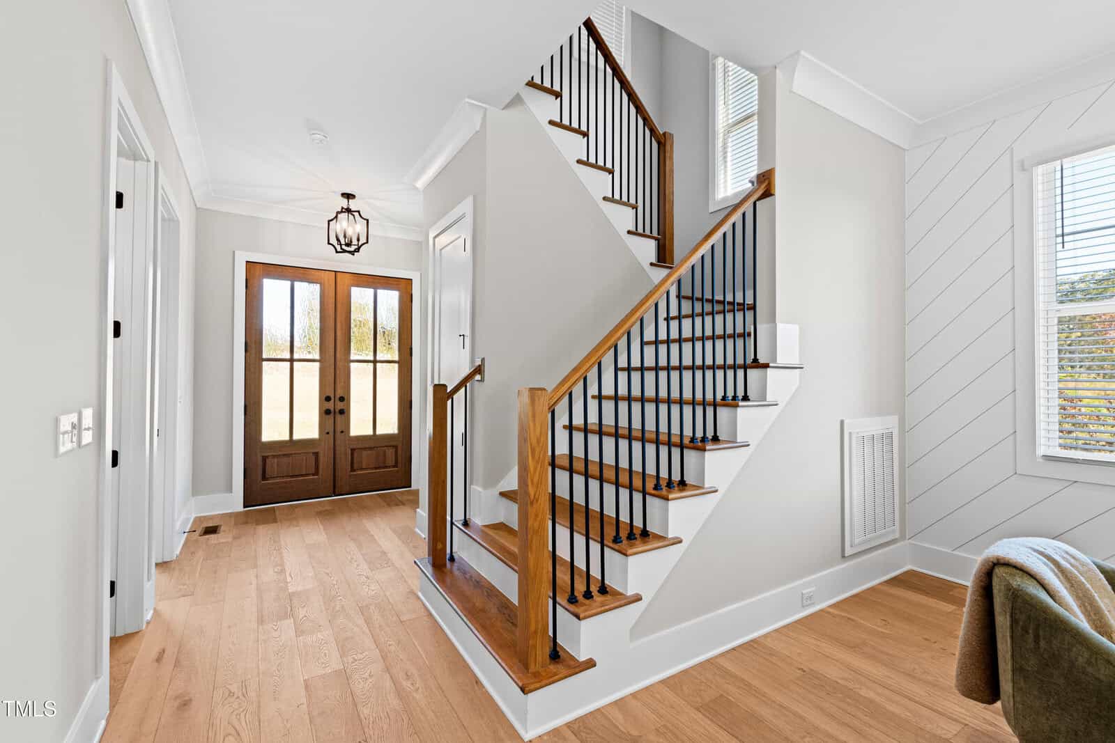 A bright entryway featuring a wooden staircase with black railing, a double front door, and light wood flooring. The walls are light-colored, and there is a window on the right side, allowing natural light to fill the space.