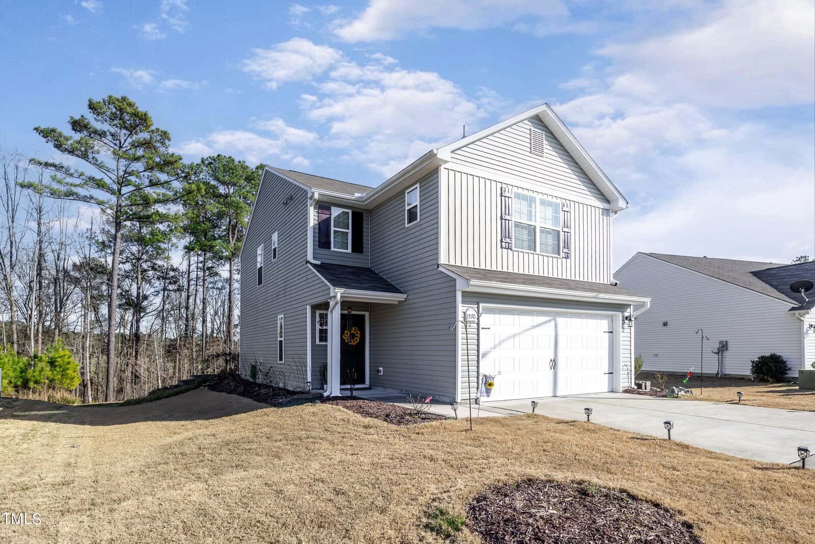 Two-story gray house with a two-car garage, set on a sloped lawn. A wreath decorates the front door. Tall trees are visible in the background under a partly cloudy sky.