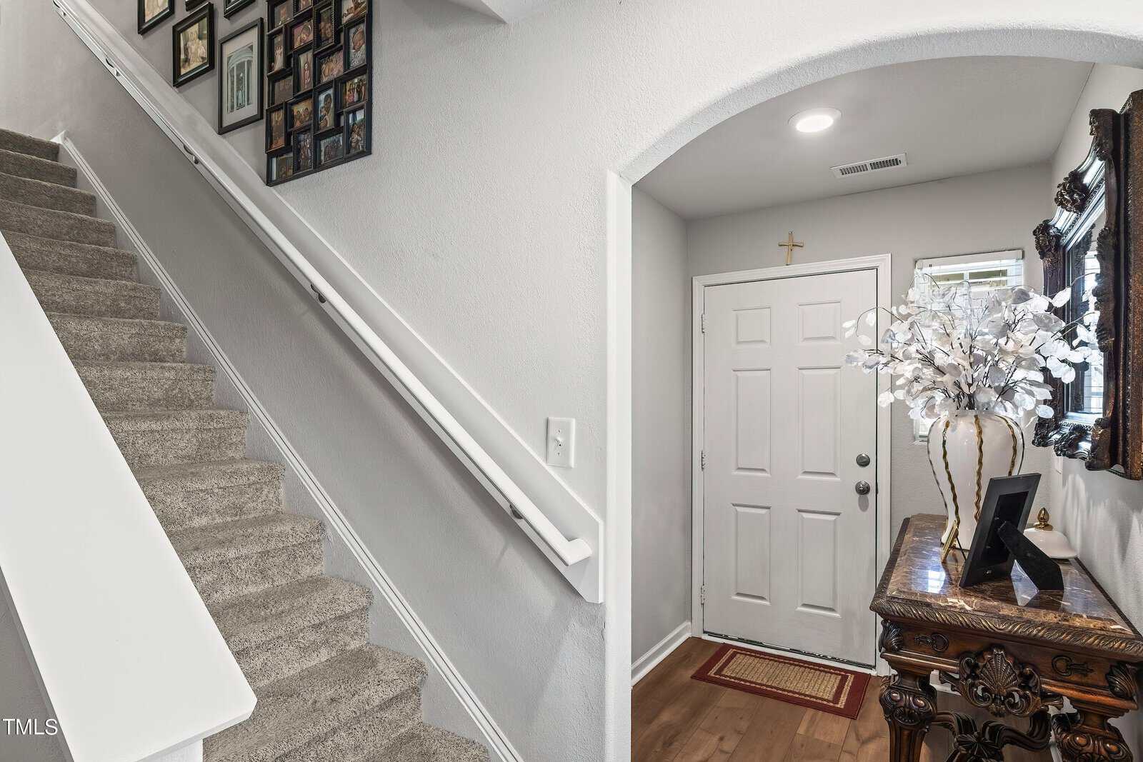 A bright entryway with a staircase on the left, leading upwards, decorated with framed photos on the wall. To the right, a wooden console table with a floral arrangement and a mirror stands against the wall by a closed white door.