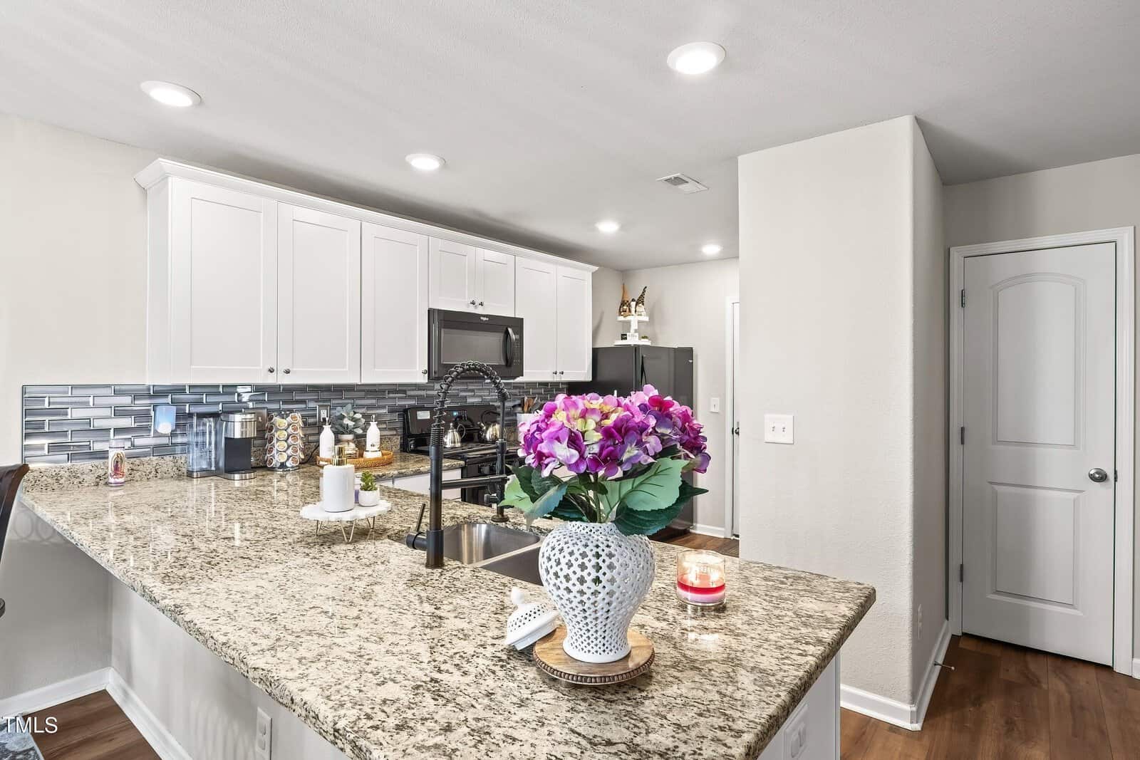 Modern kitchen with granite countertops, white cabinets, and stainless steel appliances. A bouquet of purple and pink flowers is on the island, along with a candle. The backsplash features a gray tile design. Wood flooring complements the space.