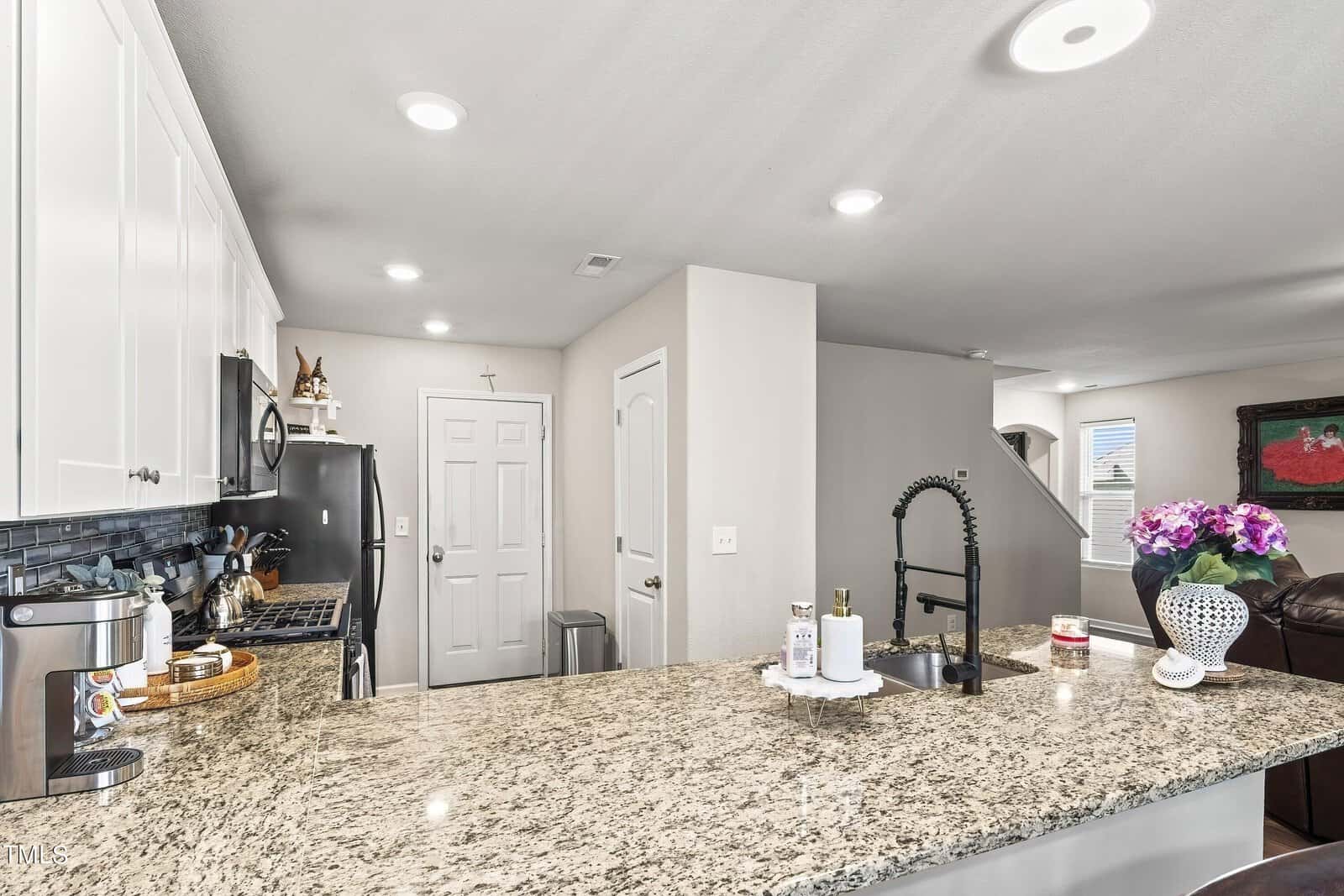 Modern kitchen with granite countertops, stainless steel appliances, and white cabinetry. A black faucet stands out on the island. In the background, there's an open living area with a brown leather couch and a vase of purple flowers.