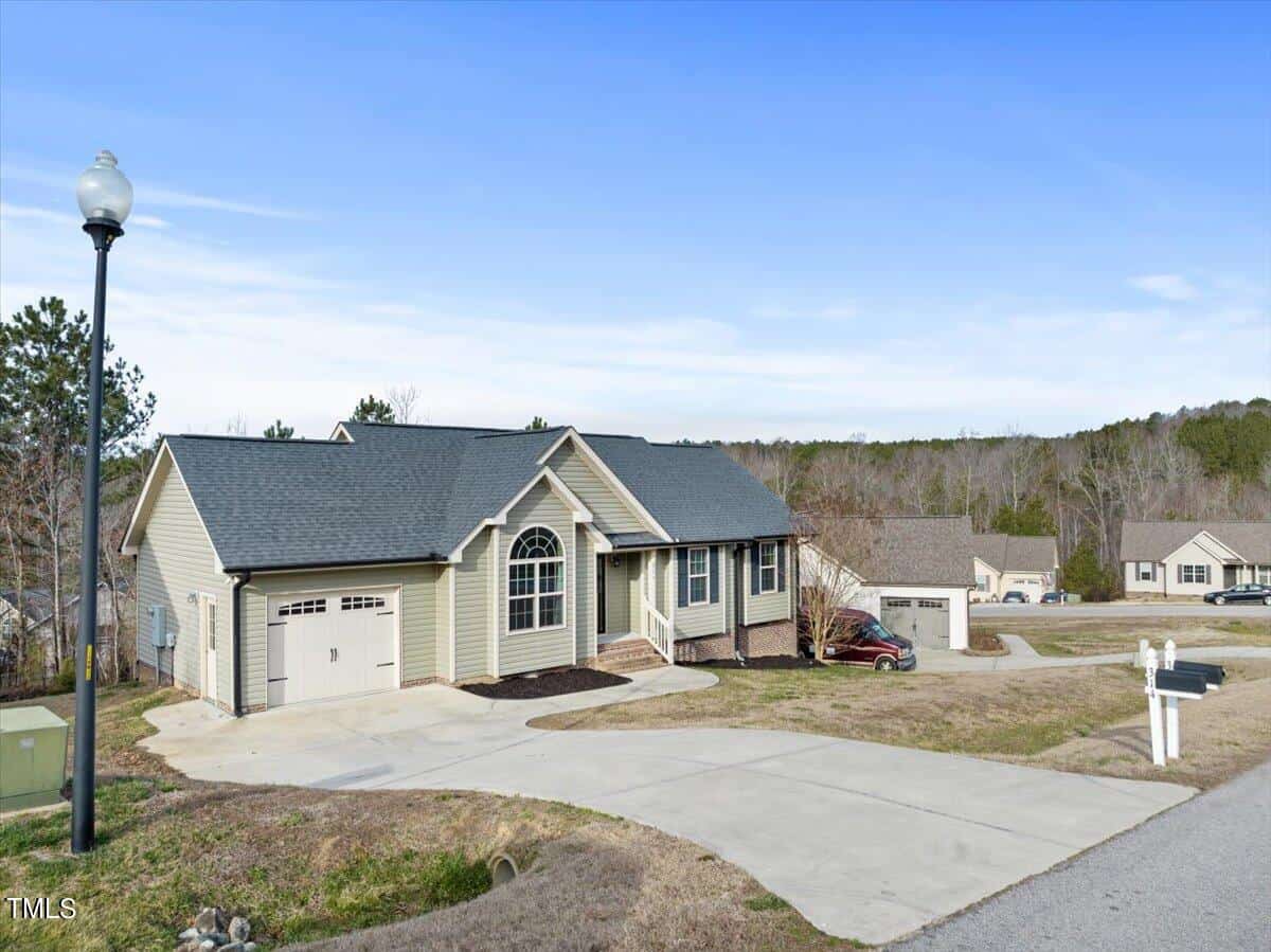 A single-story house on 315 Wick Road, with gray siding and a dark roof, sits on a sloped lawn. It features a large arched window and an attached garage. A lamppost stands nearby, casting a warm glow. Other homes stem along the street under a clear blue sky.