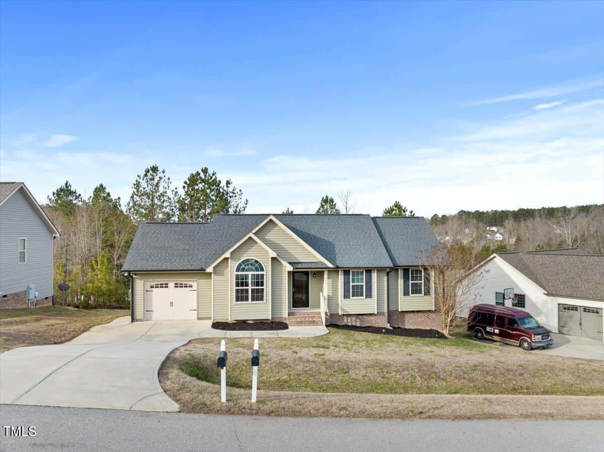 A single-story house with light-colored siding and a dark gray roof. It has a two-car garage, arched window above the entrance, and a driveway with a parked red van. The background features trees and a clear blue sky.