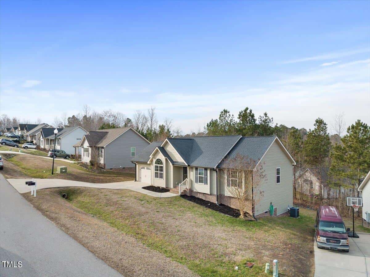 A suburban neighborhood featuring single-story houses with driveways. A light gray house with a dark roof is in the foreground on a sloped yard. A few trees and parked cars line the street under a clear blue sky.