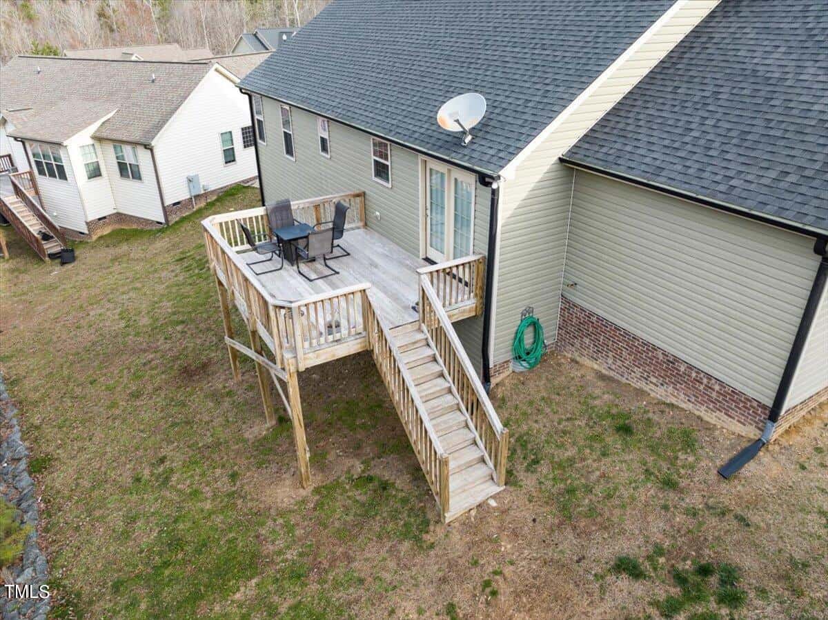 Aerial view of a house with a wooden deck featuring outdoor furniture, including a table and chairs. The deck is connected to the ground by a staircase. The house has beige siding, a gray roof, and is surrounded by a grassy yard. Adjacent structures are visible.