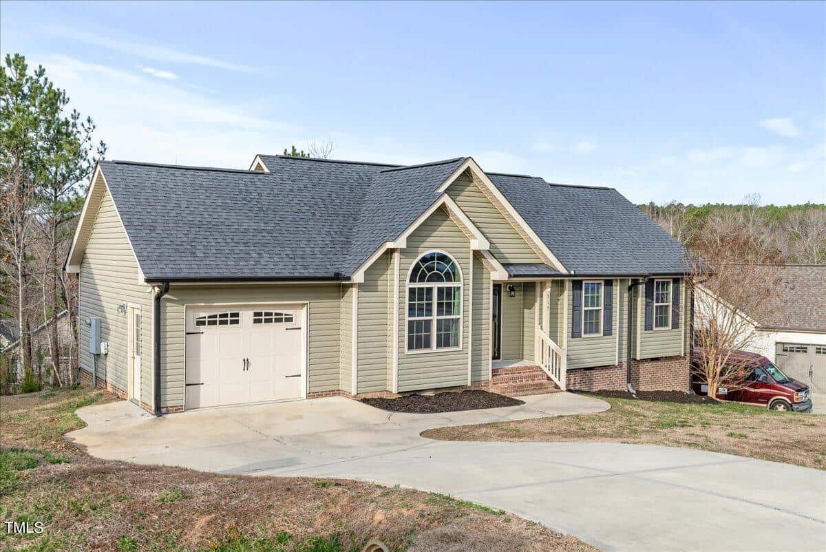 A single-story beige house with a dark gray roof and an attached garage. The front features a small porch with stairs and a large arched window. The driveway is concrete and slopes downwards. A red car is parked on the side.