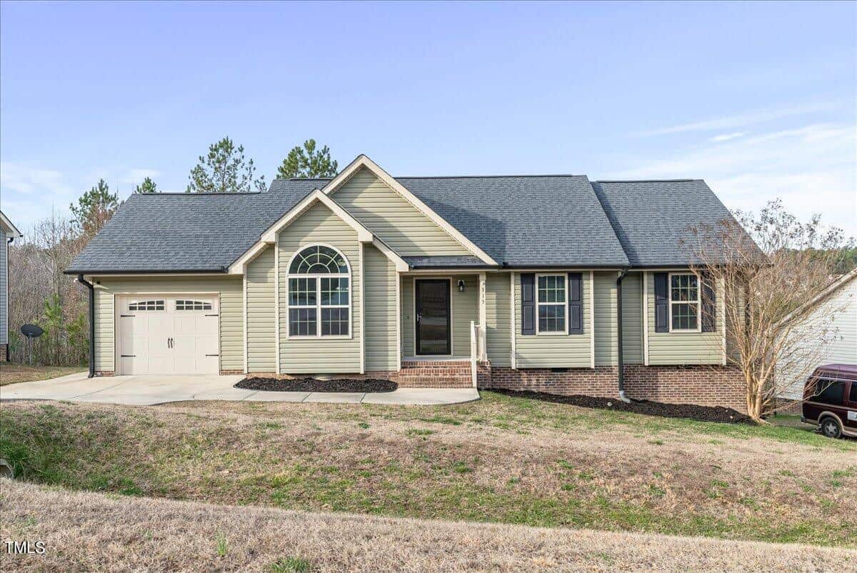 A single-story suburban house with sage green siding, white trim, and a gray roof. It features an attached garage, arched window, and a small front porch. The yard is grassy, and the sky is clear.