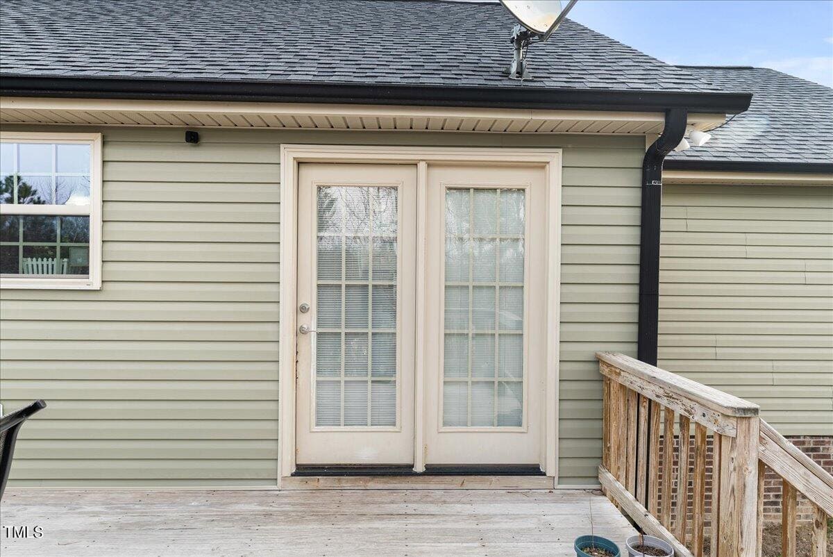 A wooden deck with French doors leading inside a house with light green siding. The doors have glass panels, and a satellite dish is attached to the roof. A small metal railing is visible on the right side.