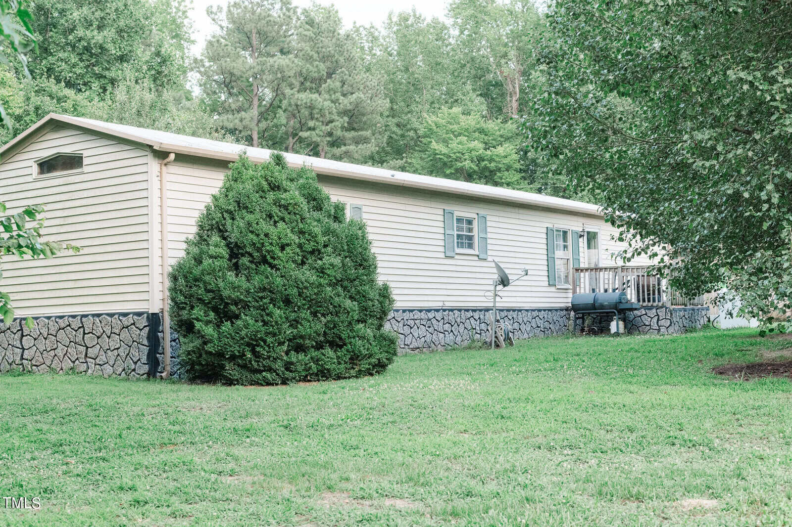 A light green mobile home on 1626 Fawn Lane boasts a wooden deck and several windows, embraced by lush greenery and trees. The stone-lined foundation complements the small satellite dish, while a well-kept lawn graces the foreground in this serene Creedmoor setting.