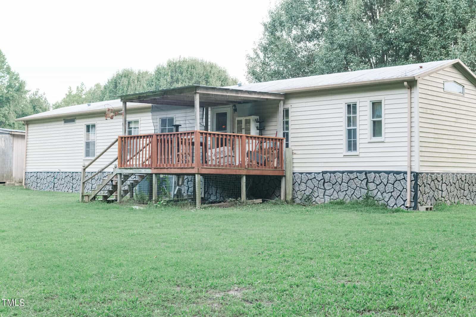 A single-story house with beige siding and a stone foundation. It features a wooden porch with chairs and railing. The house is surrounded by a grassy yard and trees in the background.