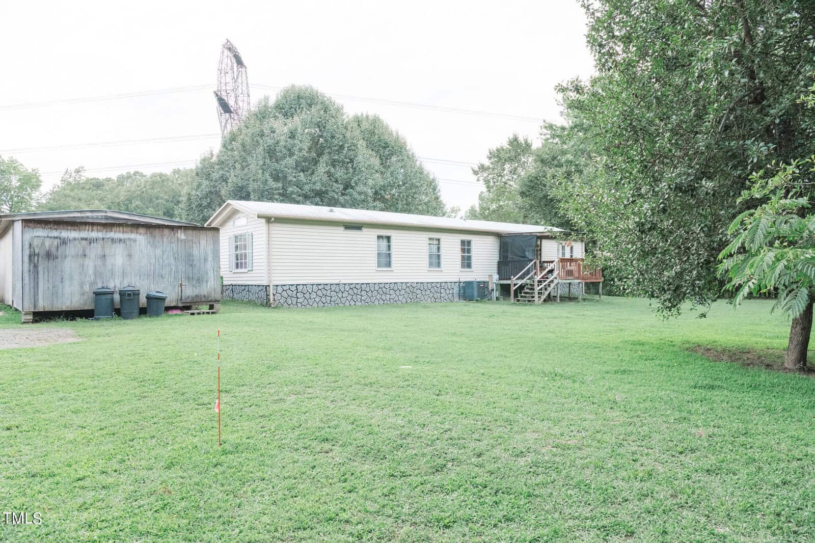 A single-story mobile home with a beige exterior and a small wooden deck is set on a large grassy yard. To the left of the home is a wooden shed, and several mature trees surround the property. A utility pole is visible in the background.