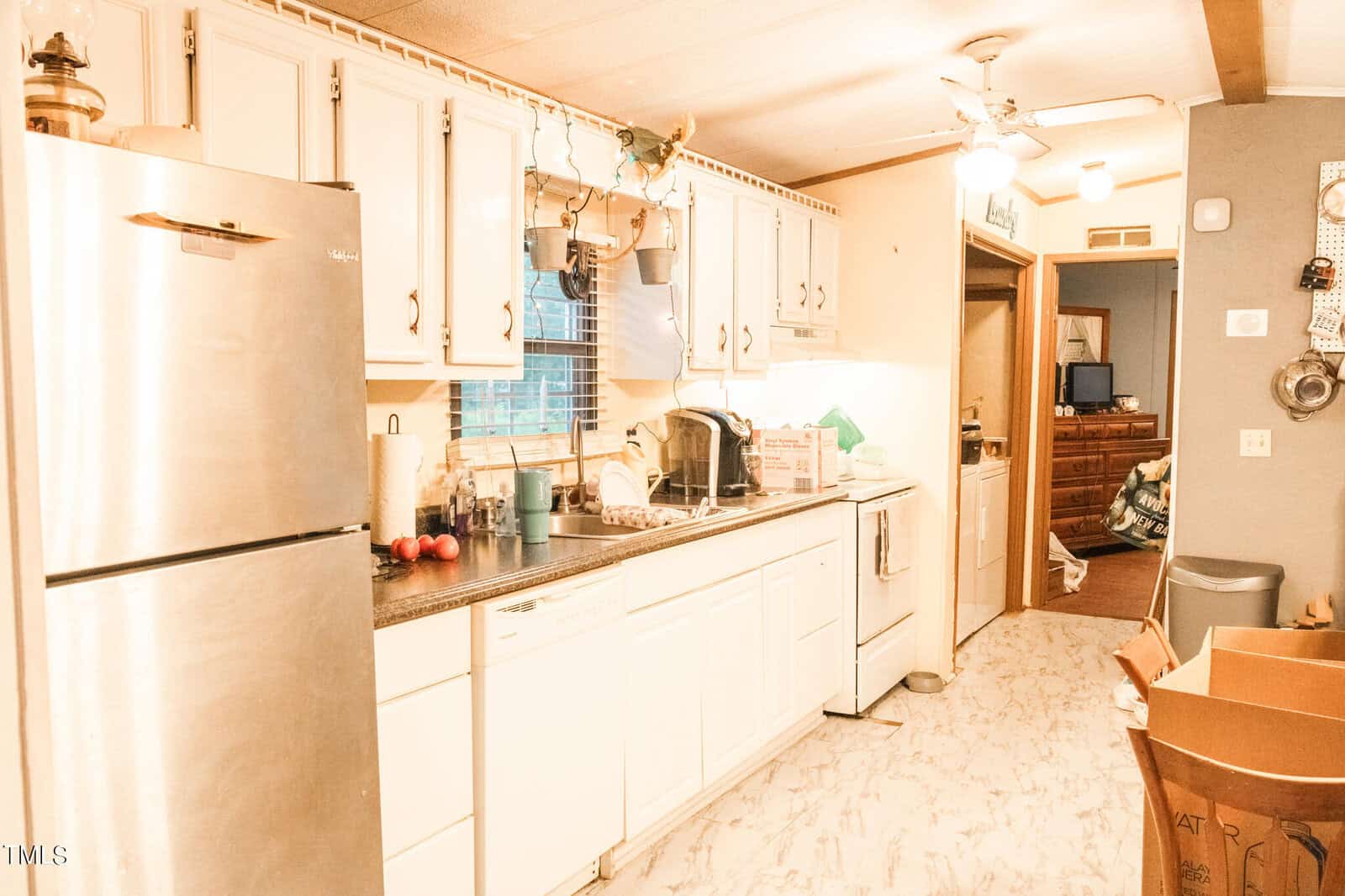 A cozy kitchen with white cabinets, a stainless steel fridge, and a countertop with various items. There's natural light coming through a window. Cardboard boxes and kitchenware are scattered around, suggesting moving or unpacking.