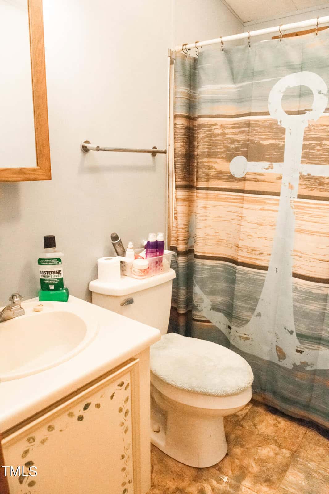 A bathroom with a sink on the left, a toilet with a cushioned lid cover, and a shower with an anchor-themed curtain. Toiletries are on the tank, including mouthwash and soap. The floor has a tile pattern, and a towel rack is on the wall.