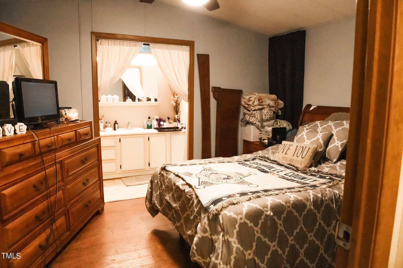 Cozy bedroom with a wooden dresser and a bed adorned with a patterned comforter. A doorway opens to a bathroom with white cabinets and a counter with toiletries. A stack of folded blankets sits on the dresser. Warm lighting fills the room.