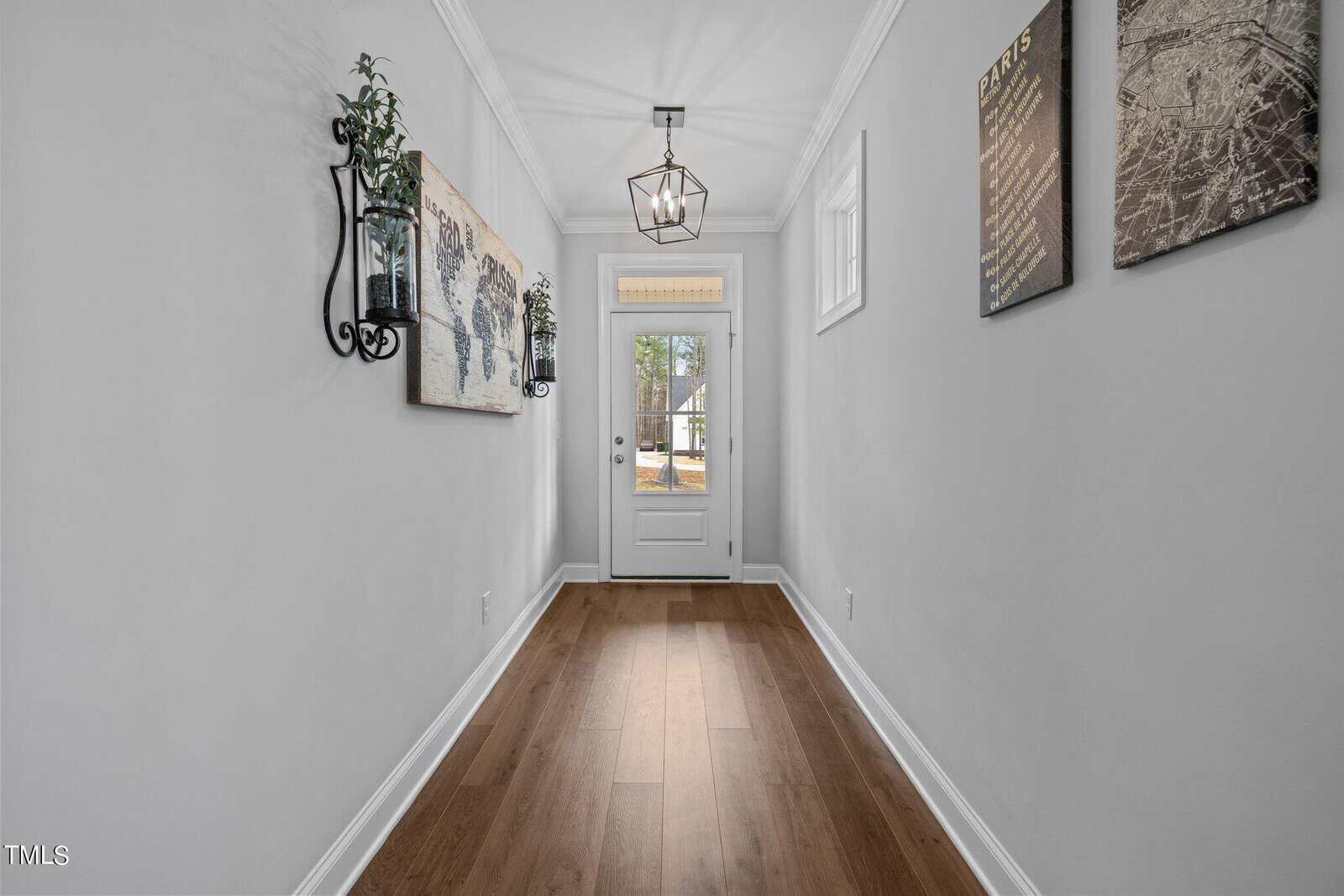 A narrow hallway with wooden floors leads to a white door with a window. Walls are decorated with a map and two framed prints. A lantern-style light hangs from the ceiling, and a potted plant is mounted on the wall. Natural light streams in.