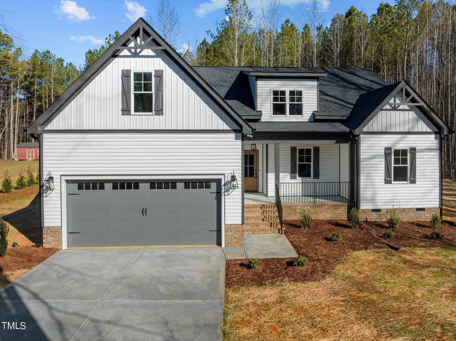 A modern, two-story house with white siding and dark gray accents. It has a two-car garage, a front porch, and surrounded by trees and a neatly landscaped yard on a sunny day.