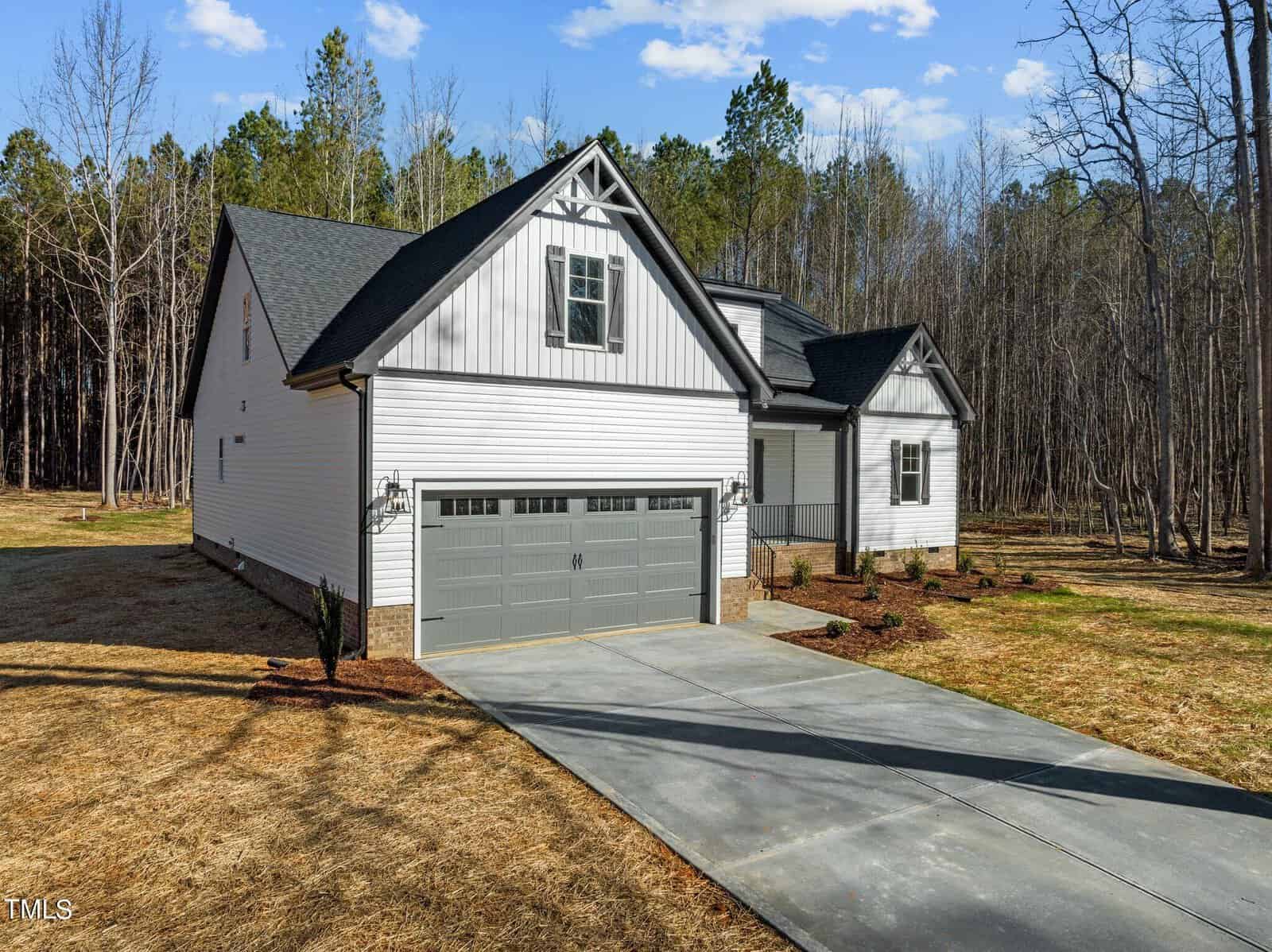 A white and gray single-story house with a gable roof and a double garage. The house is surrounded by a dry, grassy yard and bordered by a forest of tall trees under a clear blue sky.