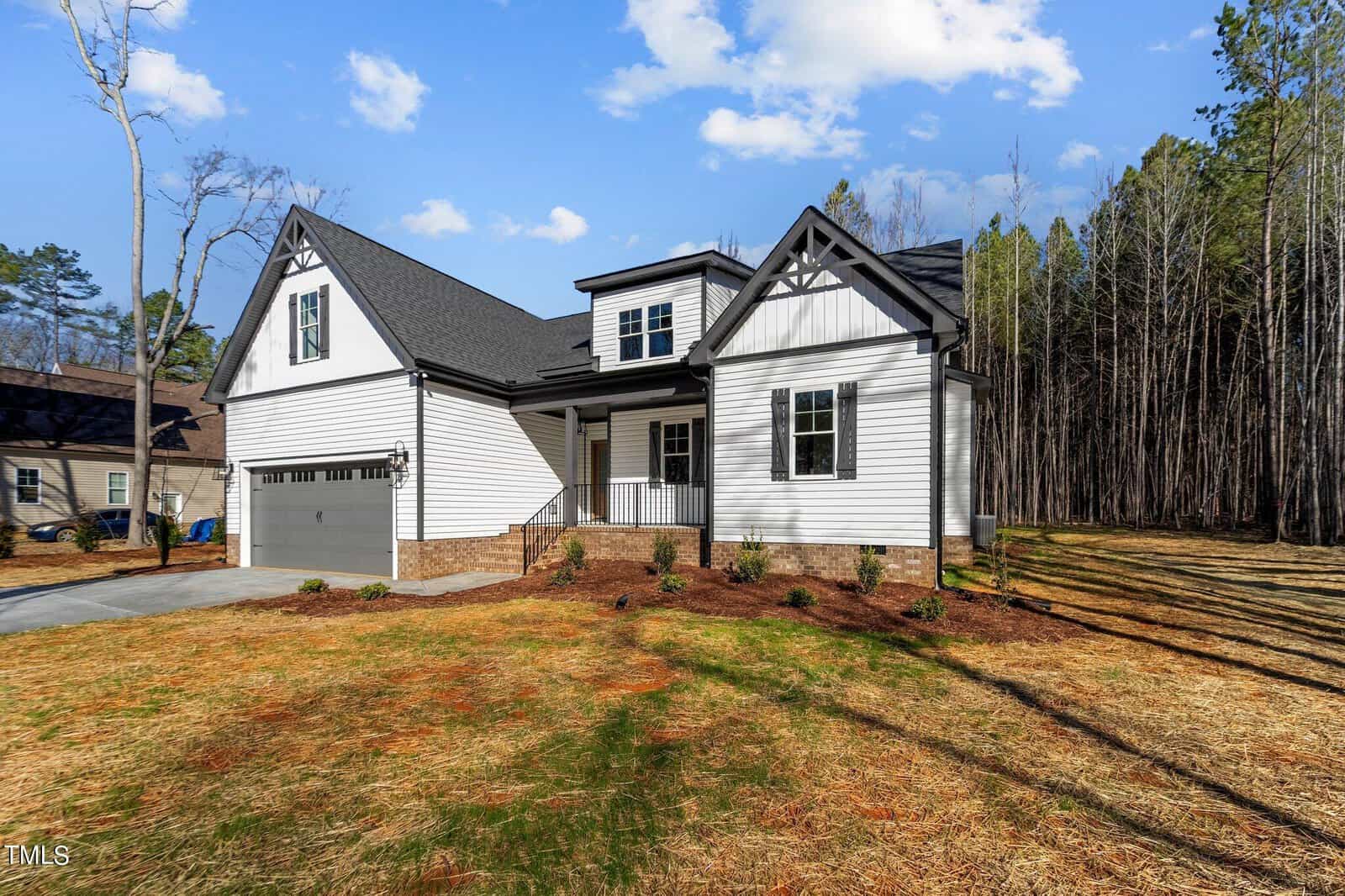 A modern two-story house with a gray roof and white siding sits surrounded by a wooded area. The driveway leads to a two-car garage. The front yard has neatly arranged brown mulch and small shrubs. The sky is partly cloudy.