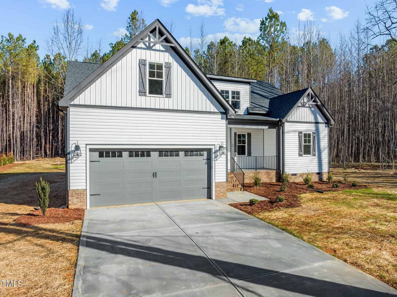 A modern, two-story white house with a gray roof and a large garage. The home features a front porch and is surrounded by a spacious lawn and tall trees. The setting is a serene, wooded area under a clear blue sky.