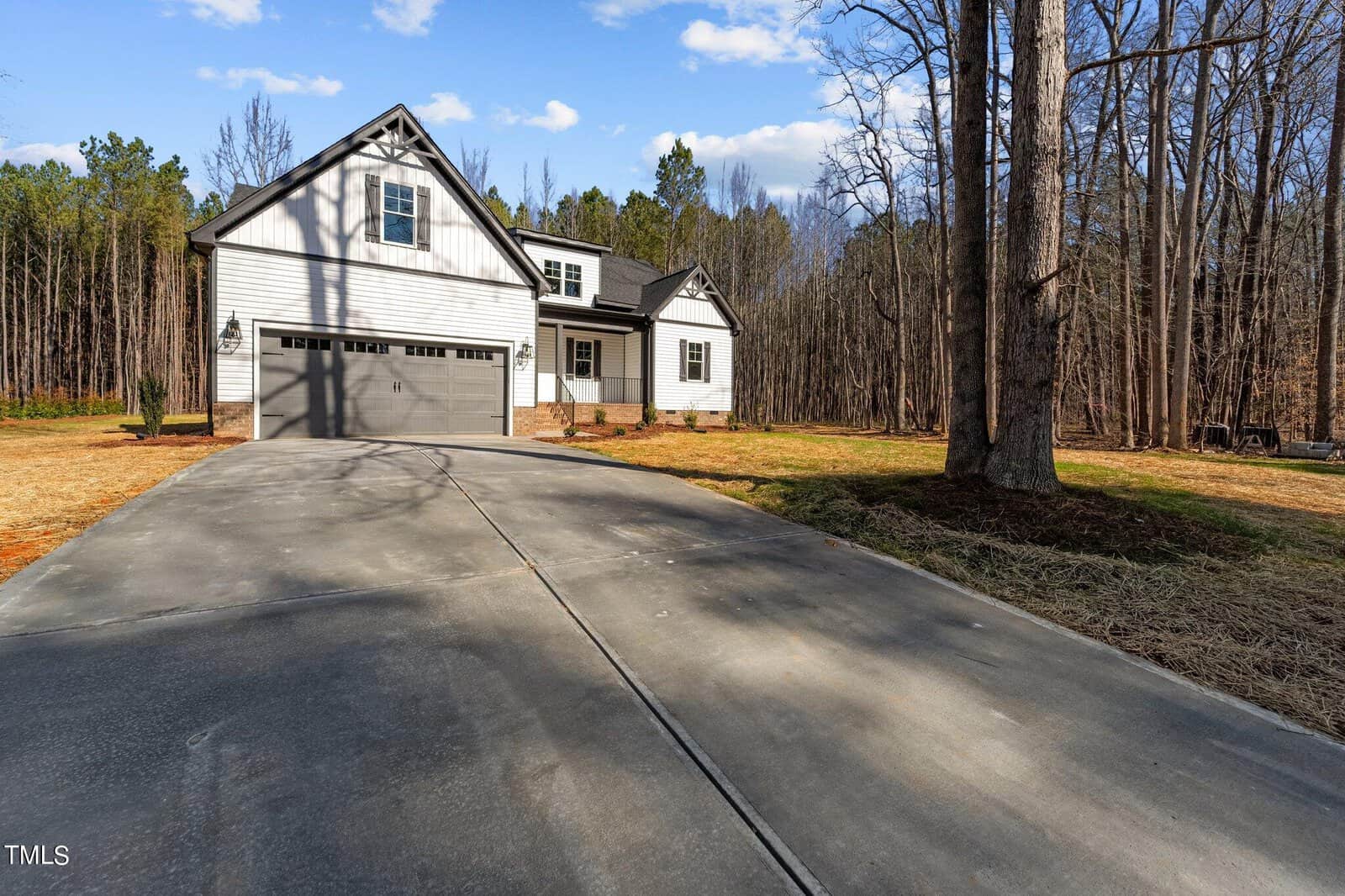 A modern white house with a two-car garage sits at the end of a long driveway surrounded by a wooded area. The sky is blue with scattered clouds, and there are tall trees with bare branches on the right side of the image.