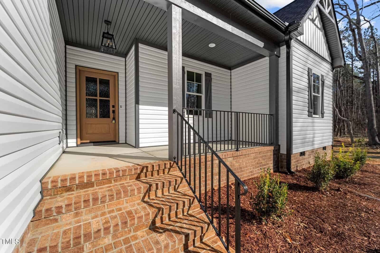 A brick house entrance with a wooden front door and black metal railing. The porch has brick steps and white siding, and it's surrounded by landscaped mulch and small green shrubs.