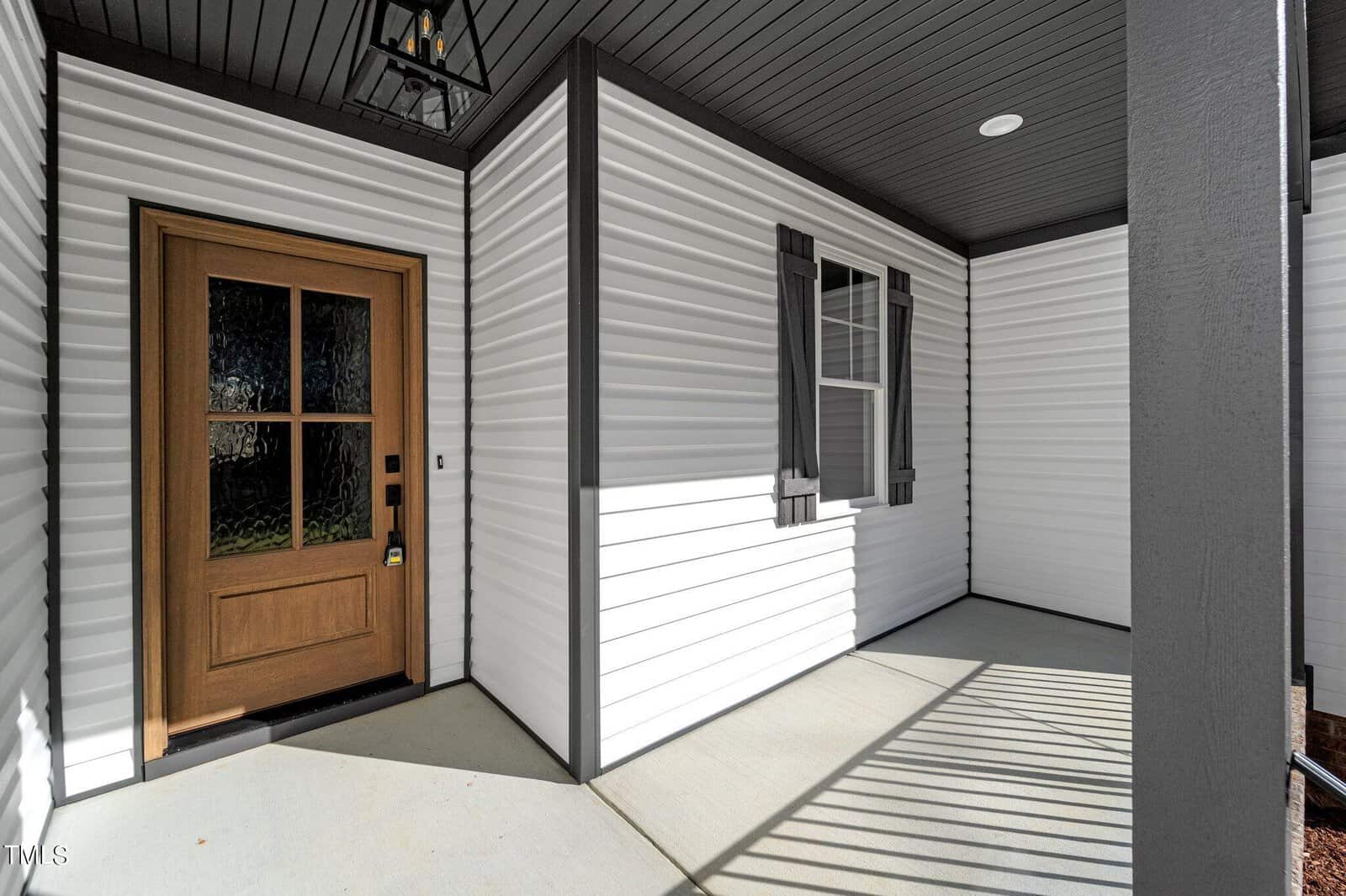 A modern, covered porch with white siding and a wooden front door featuring glass panels. A black exterior light fixture hangs overhead, and the area is accented by a window with dark shutters. Shadows from railings create patterns on the concrete floor.