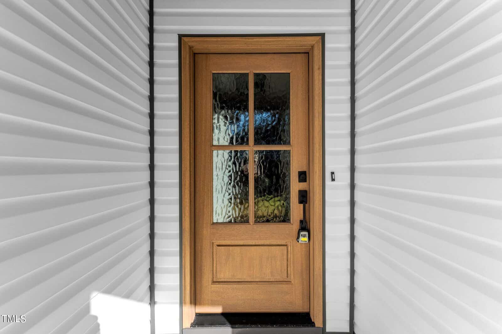 A wooden front door with a large glass panel divided into four sections. The panel has a textured pattern. The door is framed by white, ridged siding. A doorbell and lock are visible on the right side of the door.